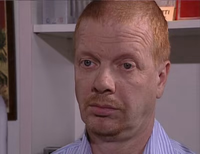 A man with red hair and a mustache, wearing a striped shirt, stares directly at the camera with a skeptical, unimpressed…