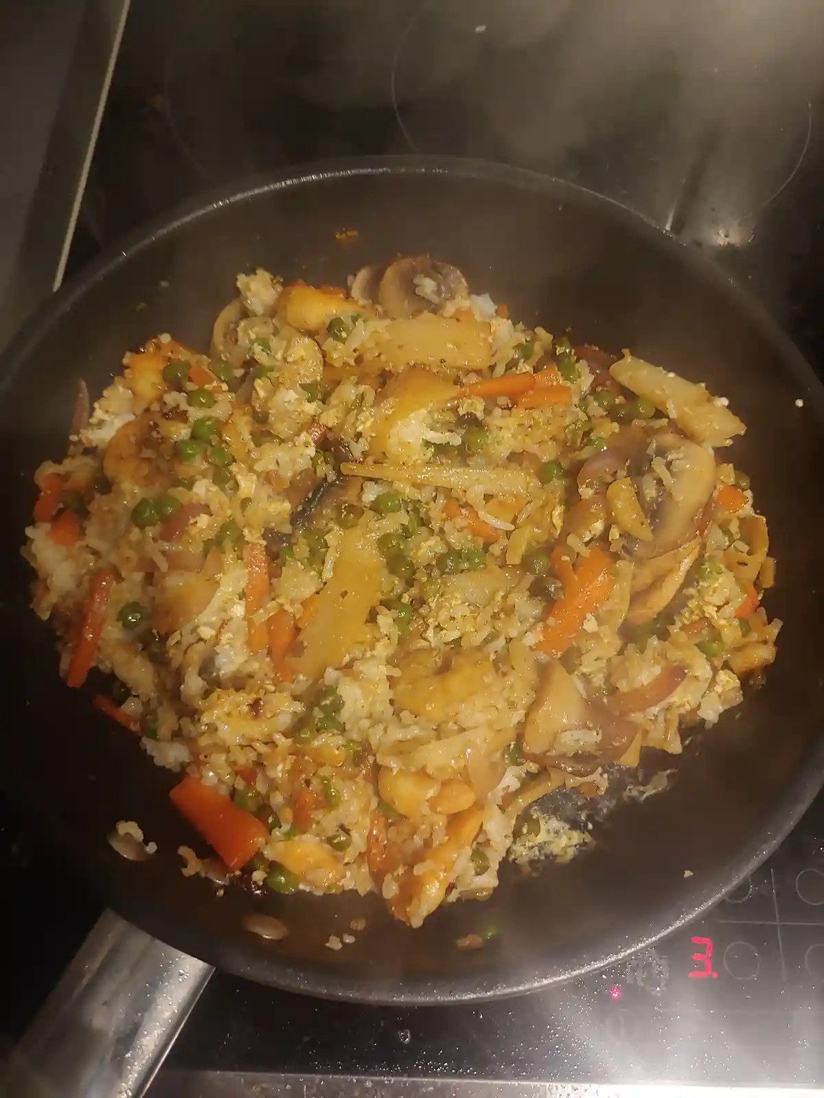 A close-up photo shows a steaming black frying pan on an electric stovetop, filled with a colorful stir-fry of rice, peas…
