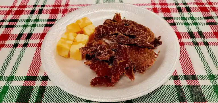 A white styrofoam plate holds a pile of beef jerky and several cubes of yellow cheese, set on a red, green, and white plaid…