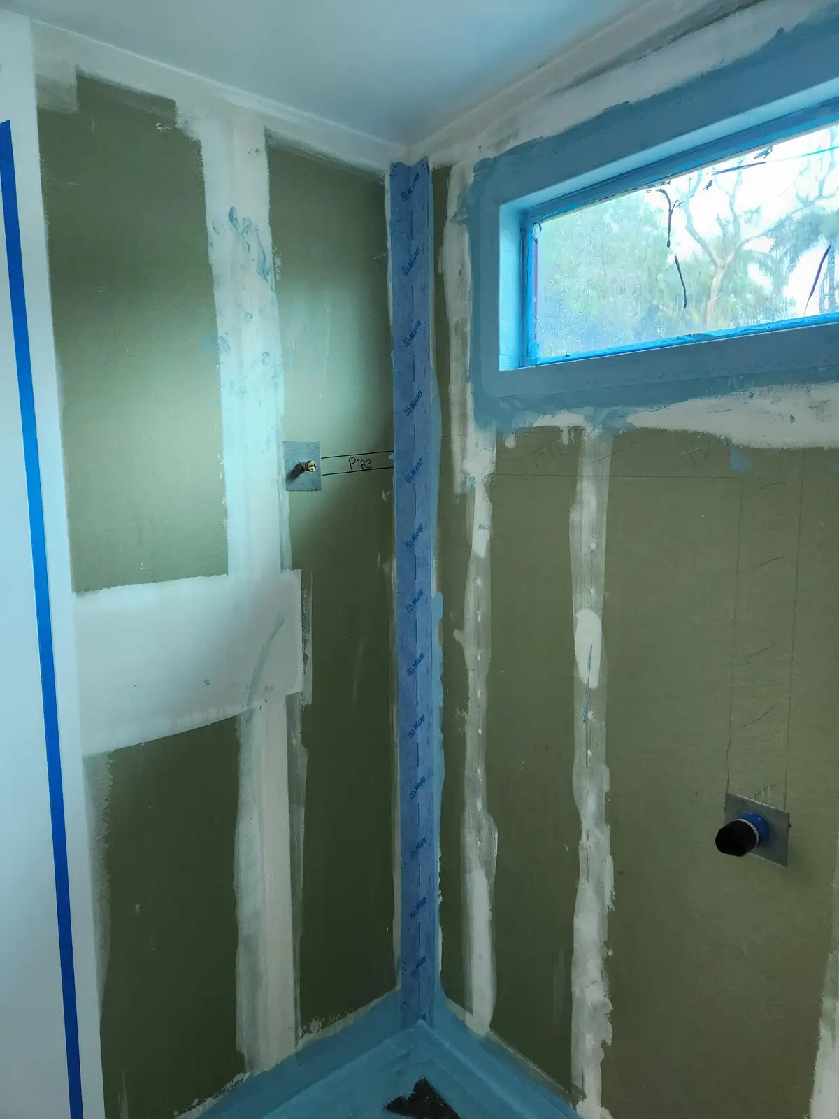 A room under renovation with walls covered in green drywall compound and blue painter's tape along edges and around a…