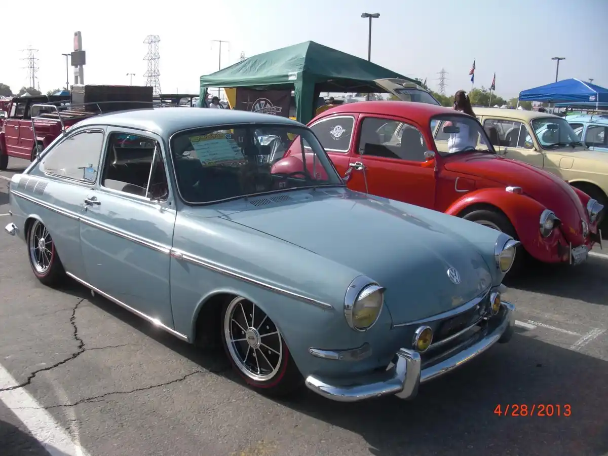 A light blue vintage Volkswagen Karmann Ghia is parked at a car show, featuring custom black wheels with red trim and chrome…