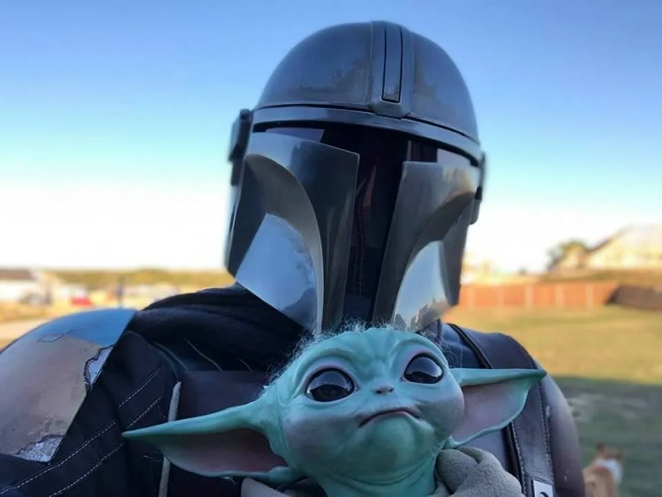 A person in a Mandalorian helmet and armor holds a small, green, child-like alien with large ears and a frowning expression…
