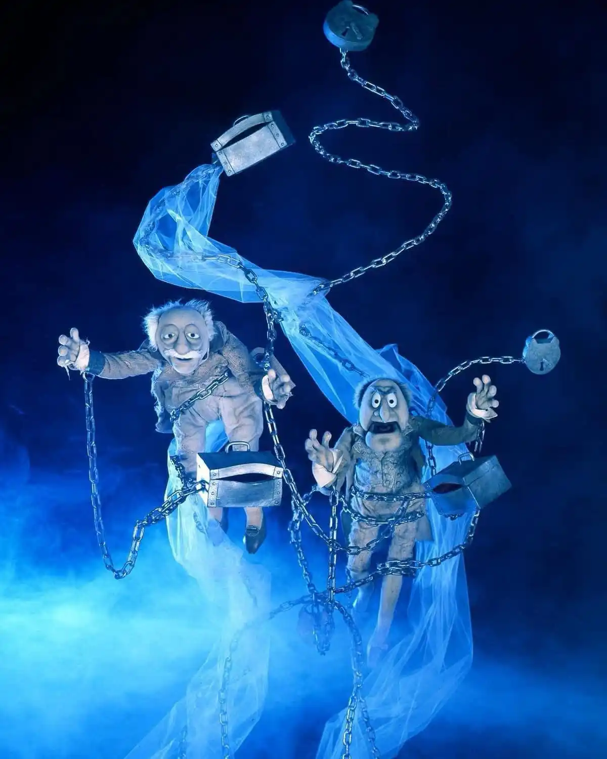 Two puppet-like figures with exaggerated, terrified expressions are suspended in mid-air by chains and padlocks, wrapped in…