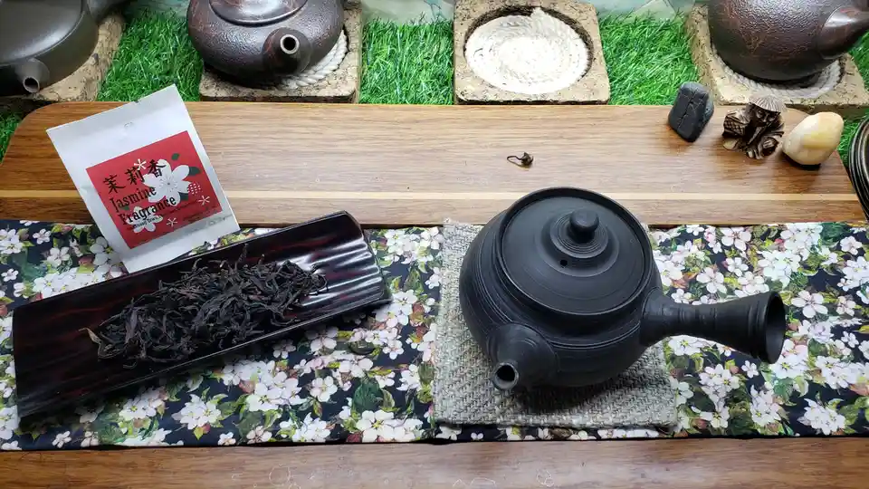 A still life of a tea setup featuring a black ceramic teapot on a woven mat atop a floral cloth, next to a wooden tray…