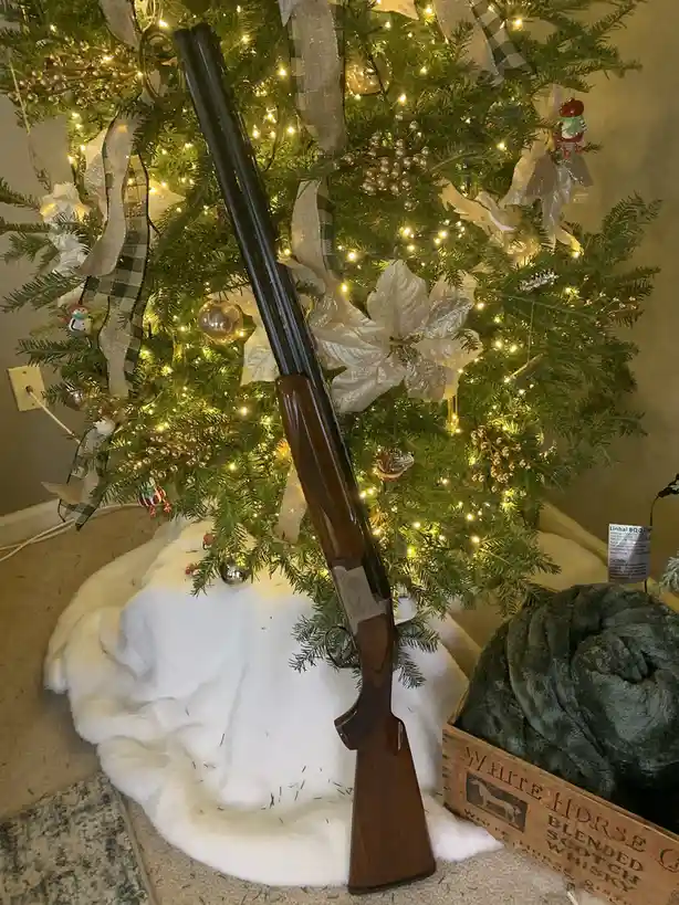 A double-barreled shotgun with a wooden stock is leaning against a decorated Christmas tree adorned with lights, ribbons…