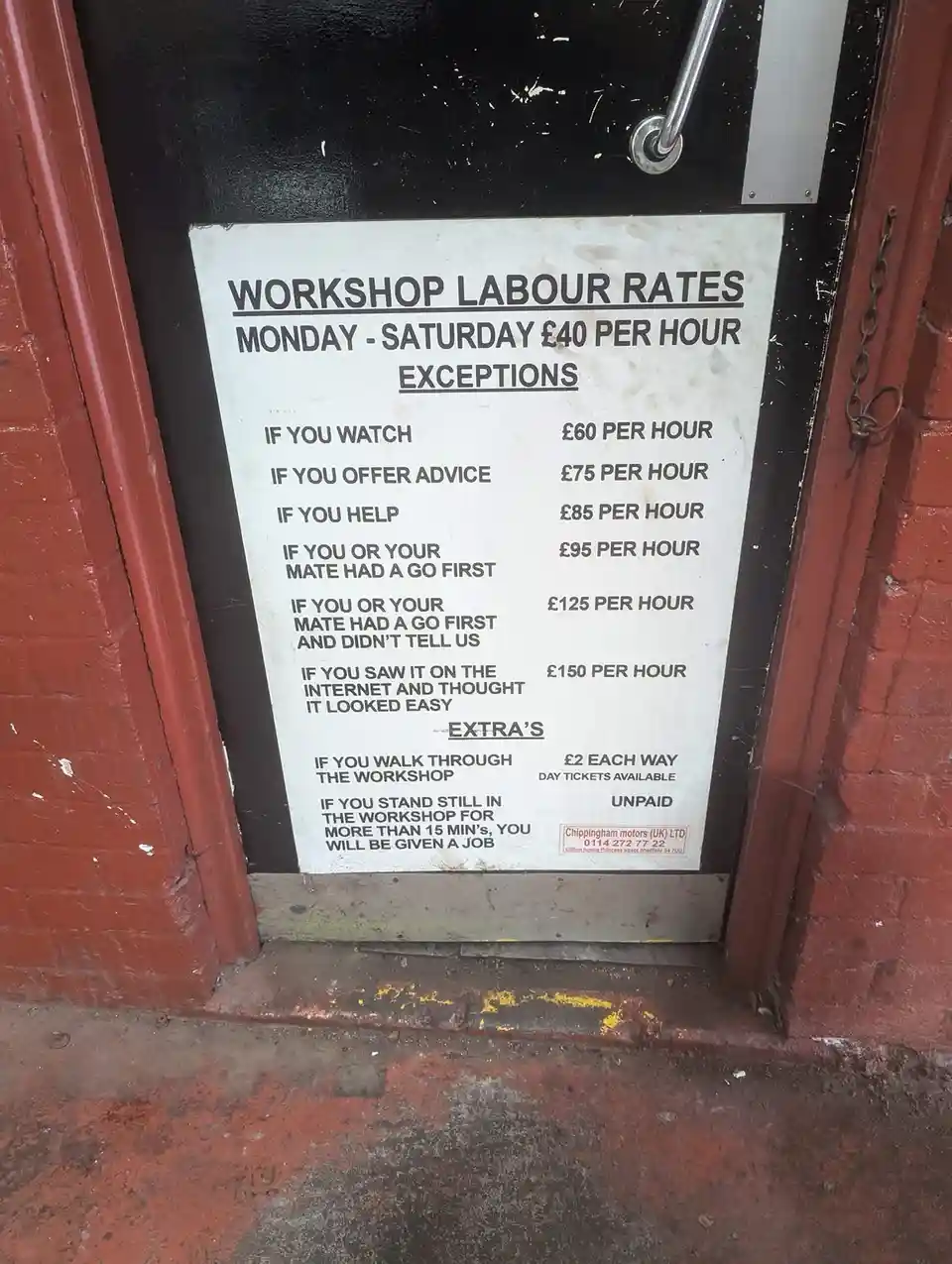 A white sign taped to a black door with red brick walls shows a satirical 'Workshop Labour Rates' list. The standard rate is…