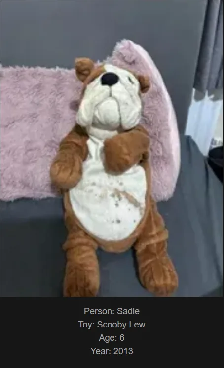 A worn, brown and white plush toy resembling a bulldog, named Scooby Lew, is shown lying on a grey couch with a pink fluffy…