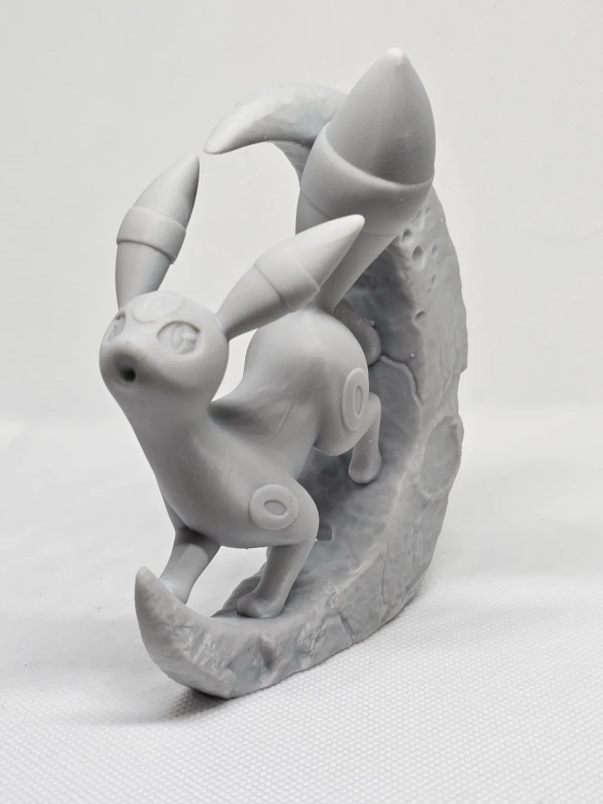 A gray, unpainted 3D-printed figurine of the Pokémon Glaceon stands on a crescent moon base, looking upward with a neutral…