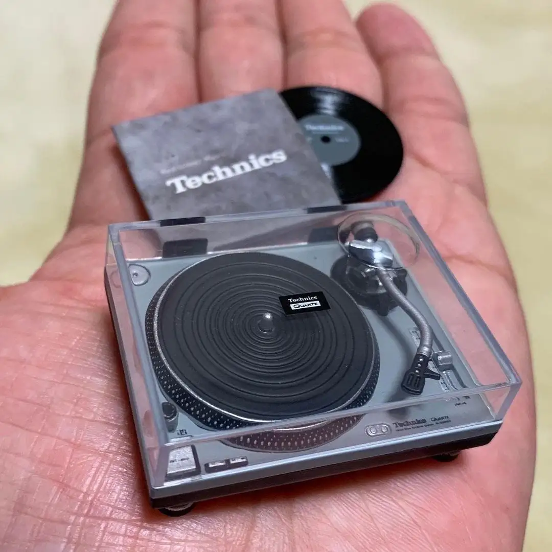 A person's open palm holds a miniature Technics turntable with a tiny vinyl record and a small grey card that reads…