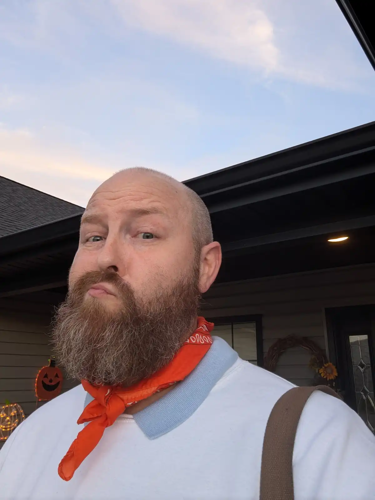 A man with a full beard and shaved head wears an orange bandana and a white shirt with a blue collar, standing outside a…