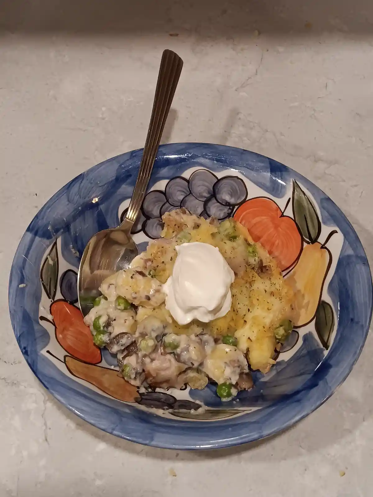 A close-up photo shows a bowl of creamy, cheesy casserole with peas and mushrooms, topped with a dollop of sour cream…