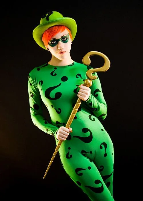 A person in a green, full-body costume covered in black question marks, wearing a matching green hat and green eye mask…