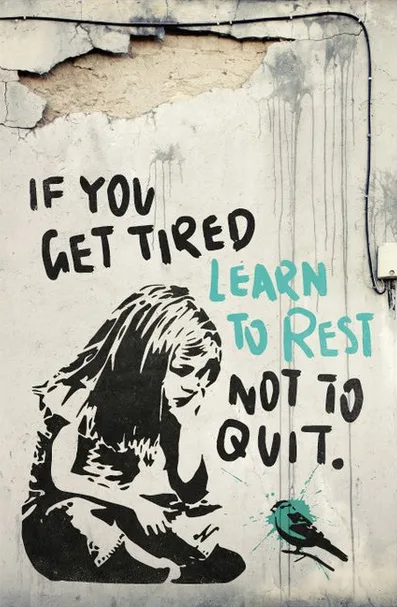 A gritty street art mural shows a black-and-white stylized woman sitting slumped with her head down, appearing exhausted…