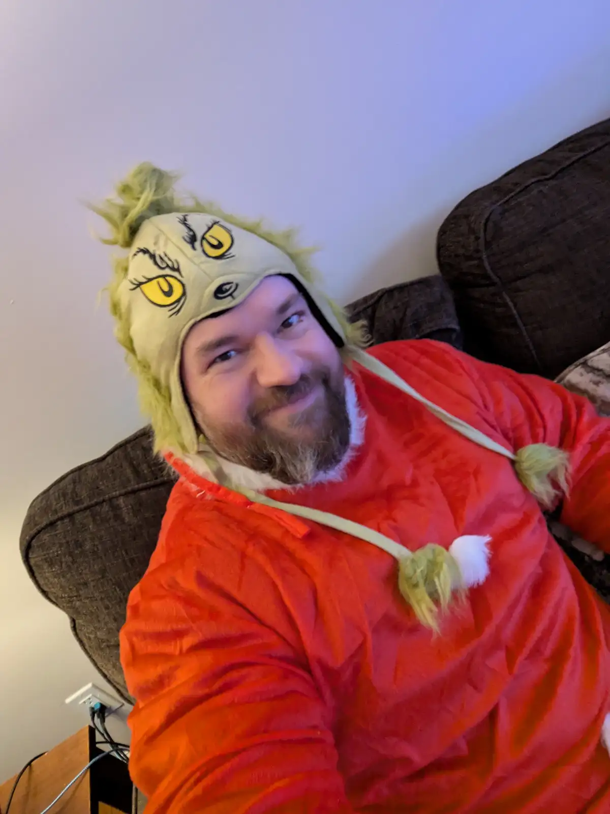 A man with a beard is lying on a couch wearing a full-body red Grinch costume with a matching hat featuring yellow eyes and…