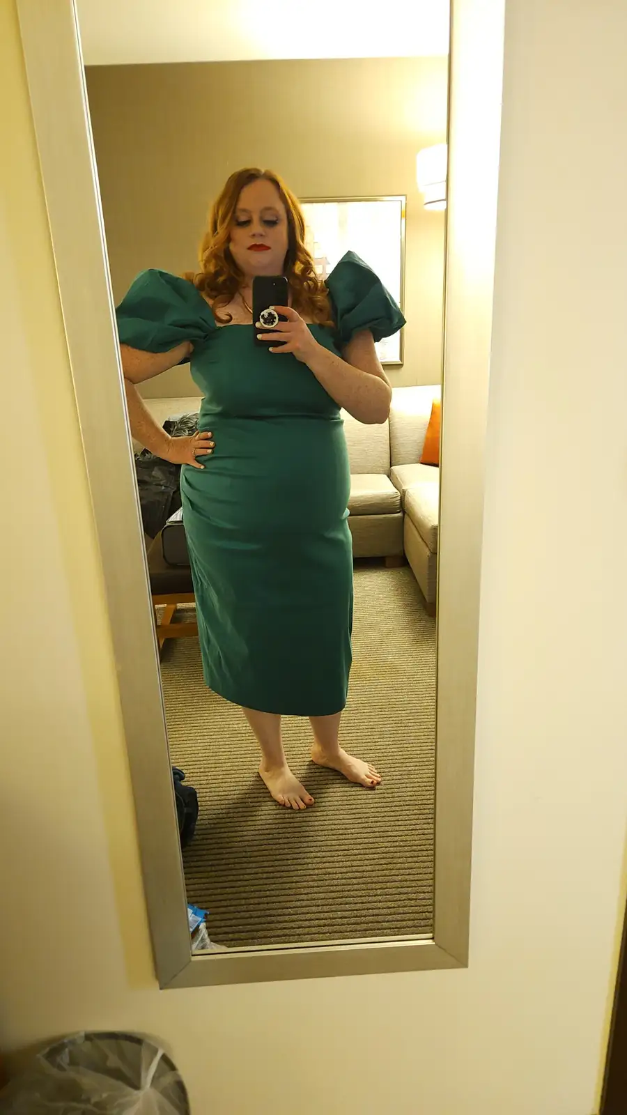 A woman with red hair and red lipstick takes a mirror selfie in a hotel room, wearing a form-fitting teal dress with puffy…