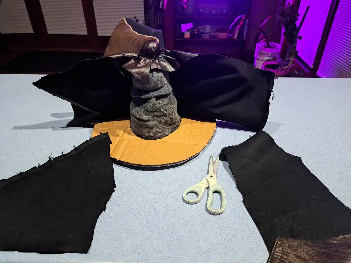 A handmade witch hat sits on a table, constructed from a cardboard base, a gray sock forming the cone, and black fabric for…