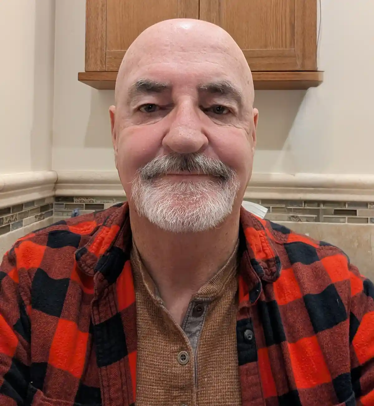 A bald man with a white goatee and mustache, wearing a red and black plaid flannel shirt over a brown button-up, looks…