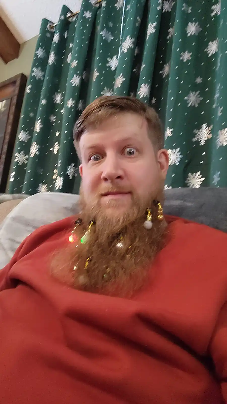 A man with a long brown beard sits on a couch wearing a red sweater, with a string of lit Christmas lights wrapped around…