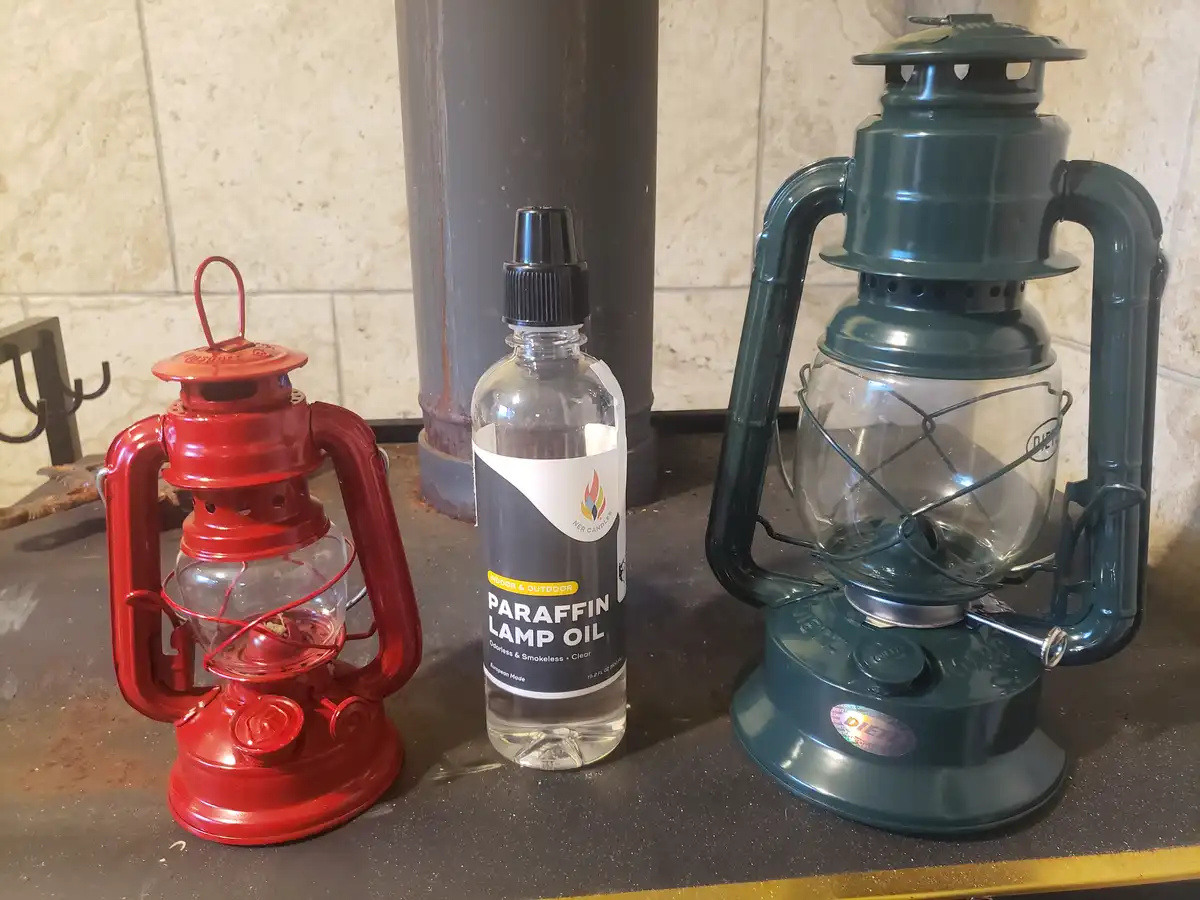 A red and a green kerosene-style lantern sit on a dark countertop beside a clear bottle labeled 'PARAFFIN LAMP OIL'. The…