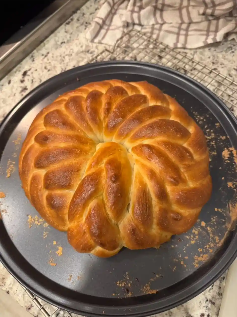 A golden-brown braided bread with a flower-like pattern sits on a black baking pan, resting on a wire cooling rack atop a…