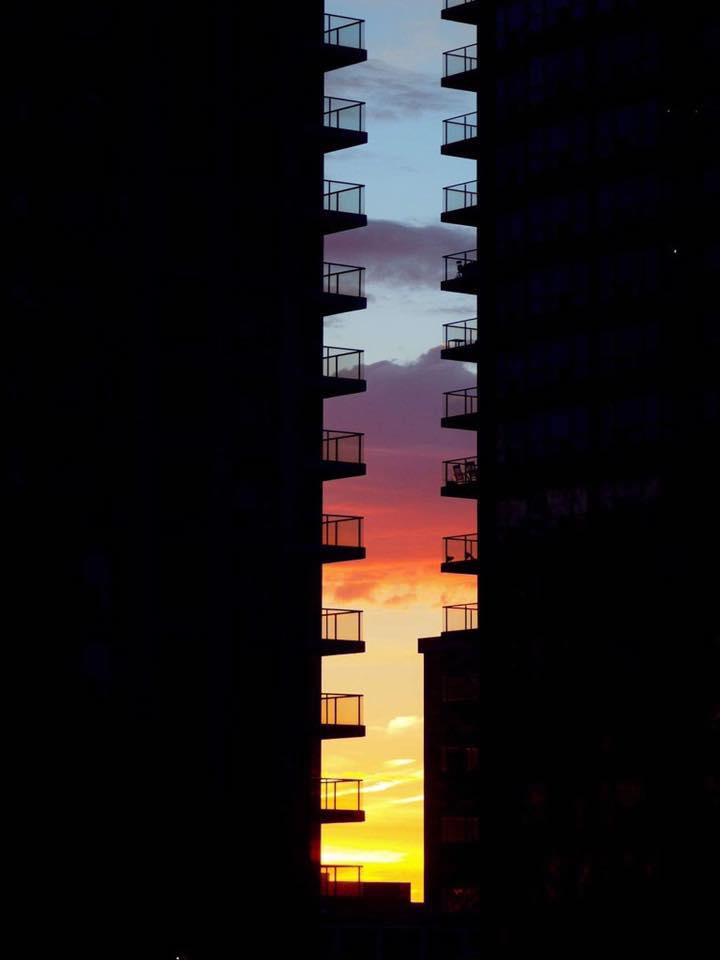 A photograph shows the silhouette of two tall apartment buildings framing a vibrant sunset with orange, pink, and blue hues…