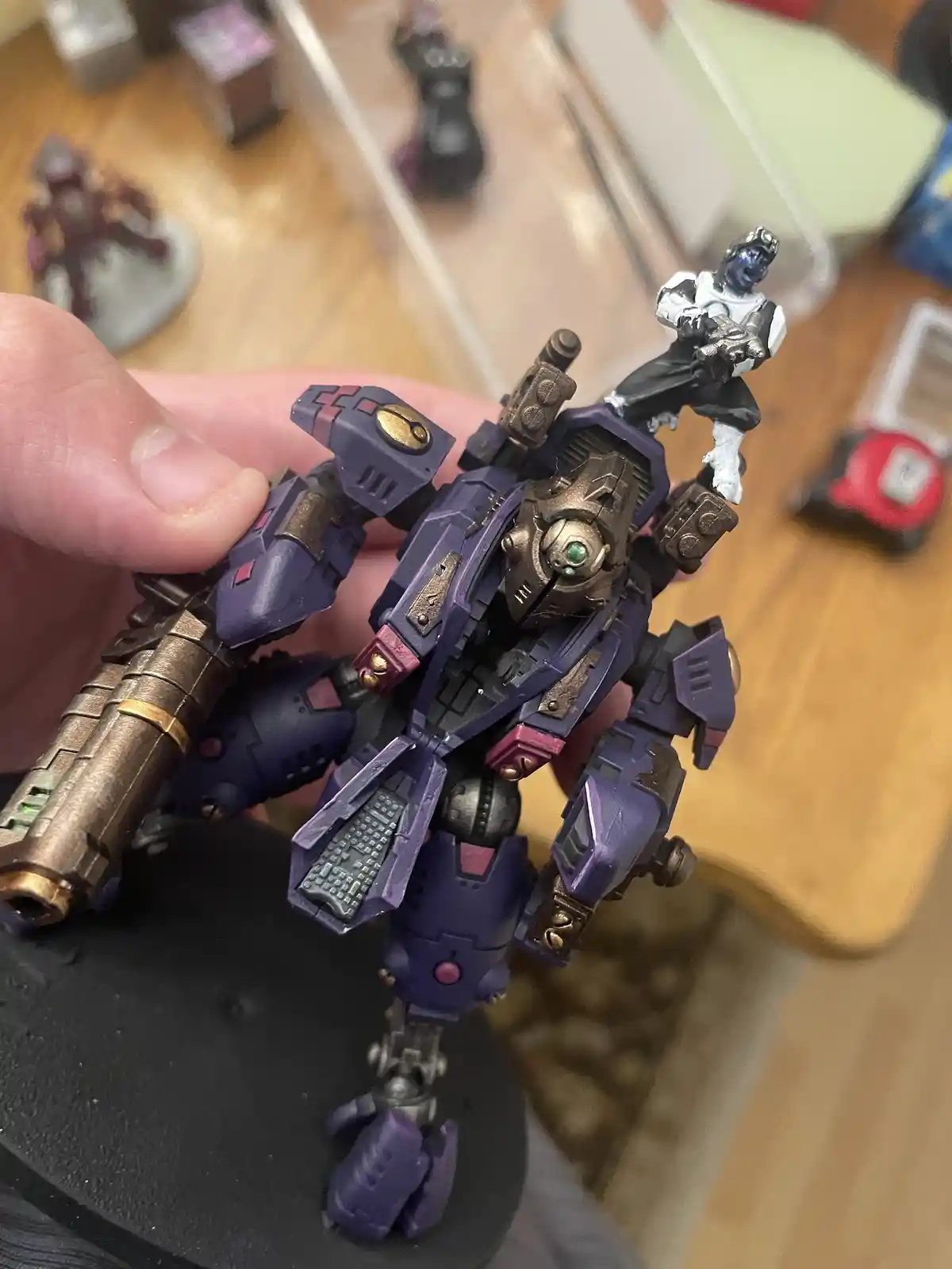 A hand holds a detailed purple and gold painted miniature robot with a small blue-skinned humanoid figure perched on its…