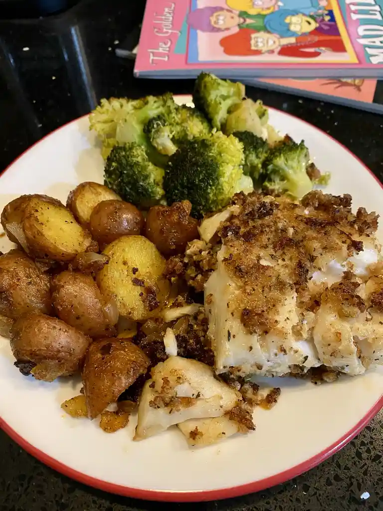 A plate of cooked food including roasted potatoes, broccoli, and breaded fish is shown on a black countertop. In the…