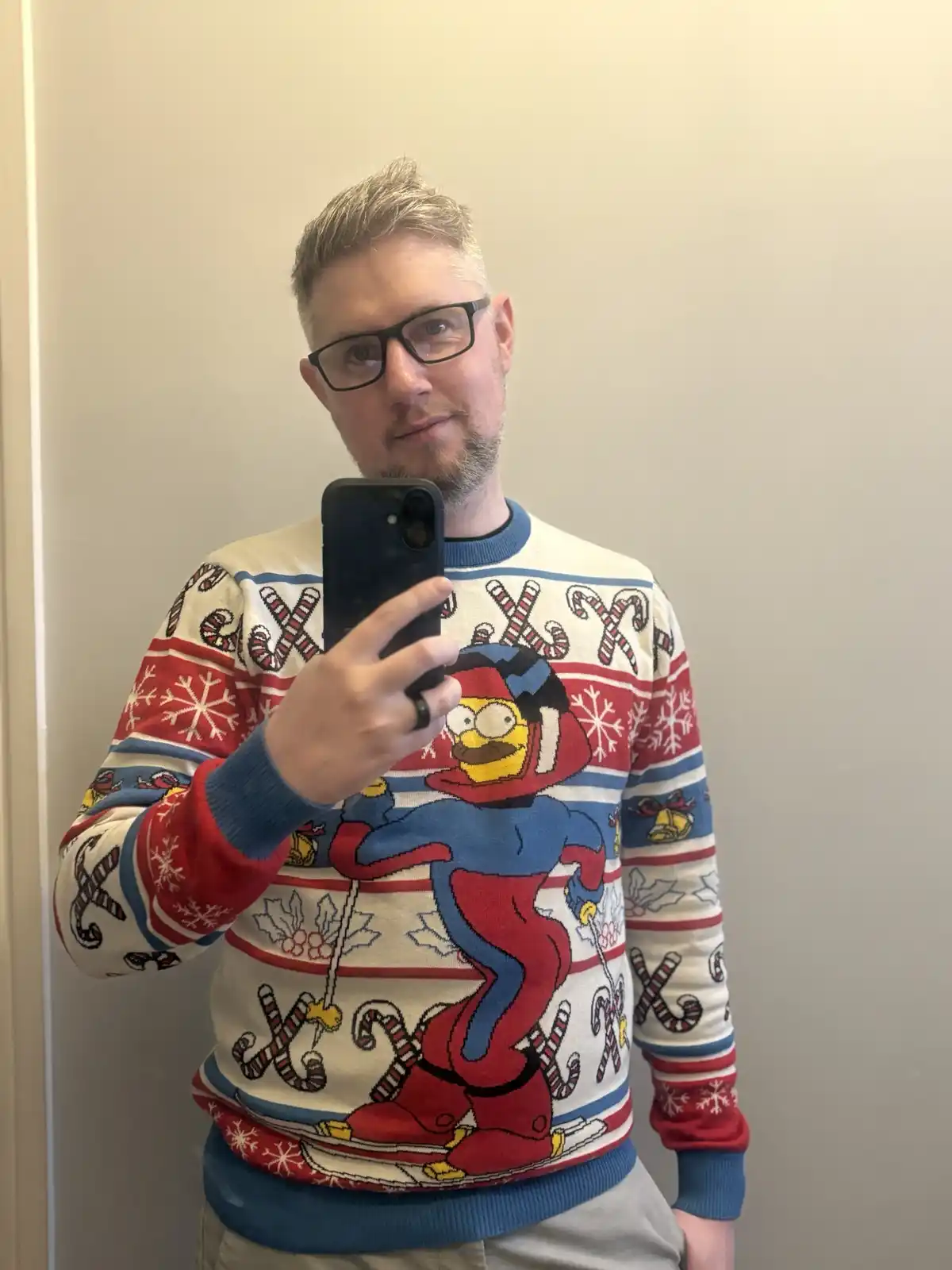A man with glasses and a beard takes a mirror selfie wearing a very busy, colorful Christmas sweater featuring Homer Simpson…