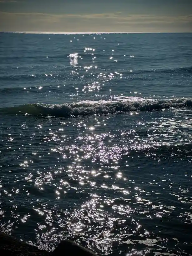 A serene photograph of ocean waves with sunlight glinting off the water's surface, creating sparkling reflections. The…