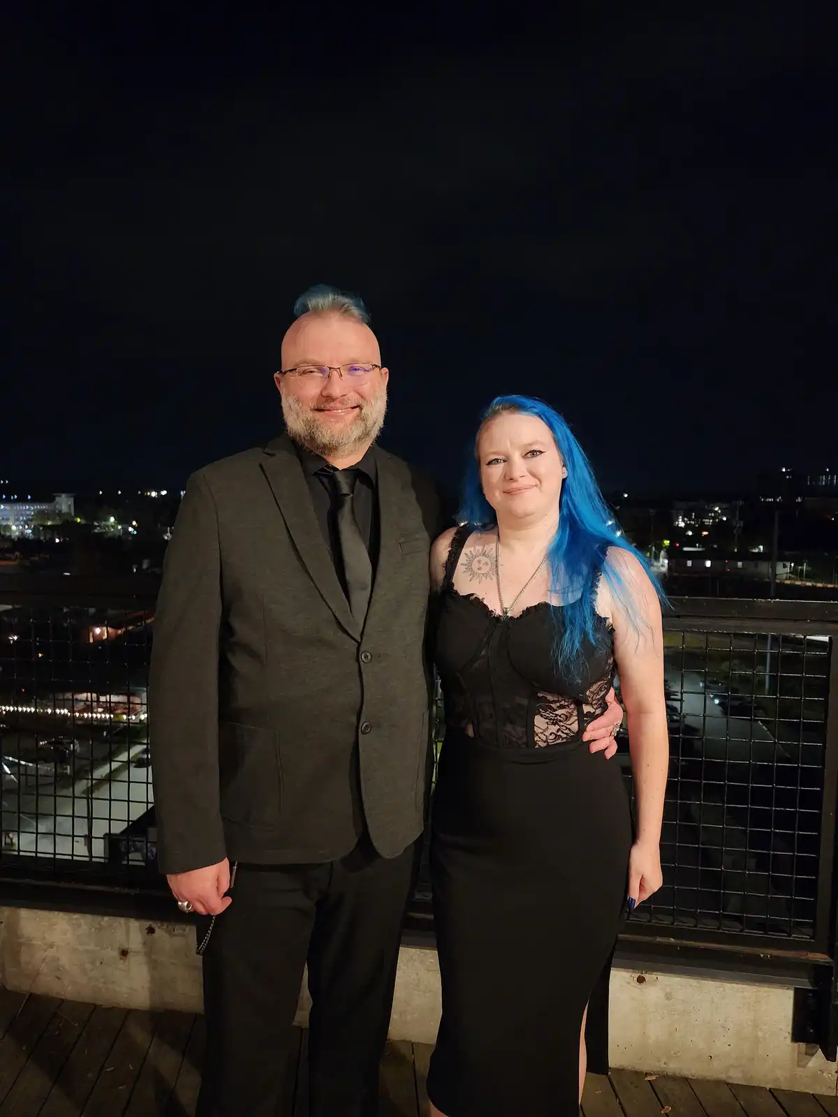 A man and woman pose together on a rooftop at night, both with blue hair, the man has a small blue mohawk and the woman has…