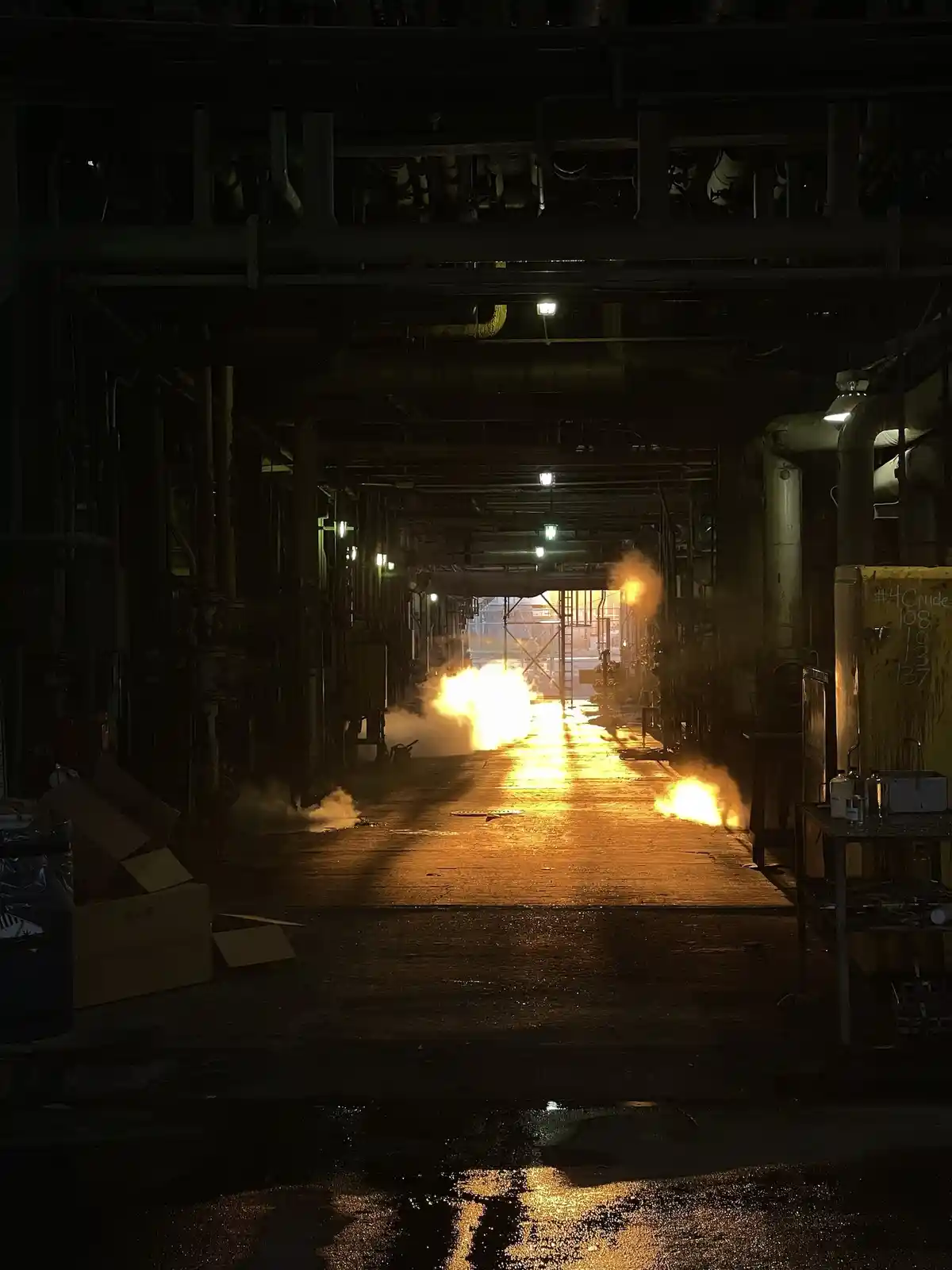 A dimly lit industrial corridor with pipes overhead and a wet floor, where bright orange flames and smoke erupt from the…