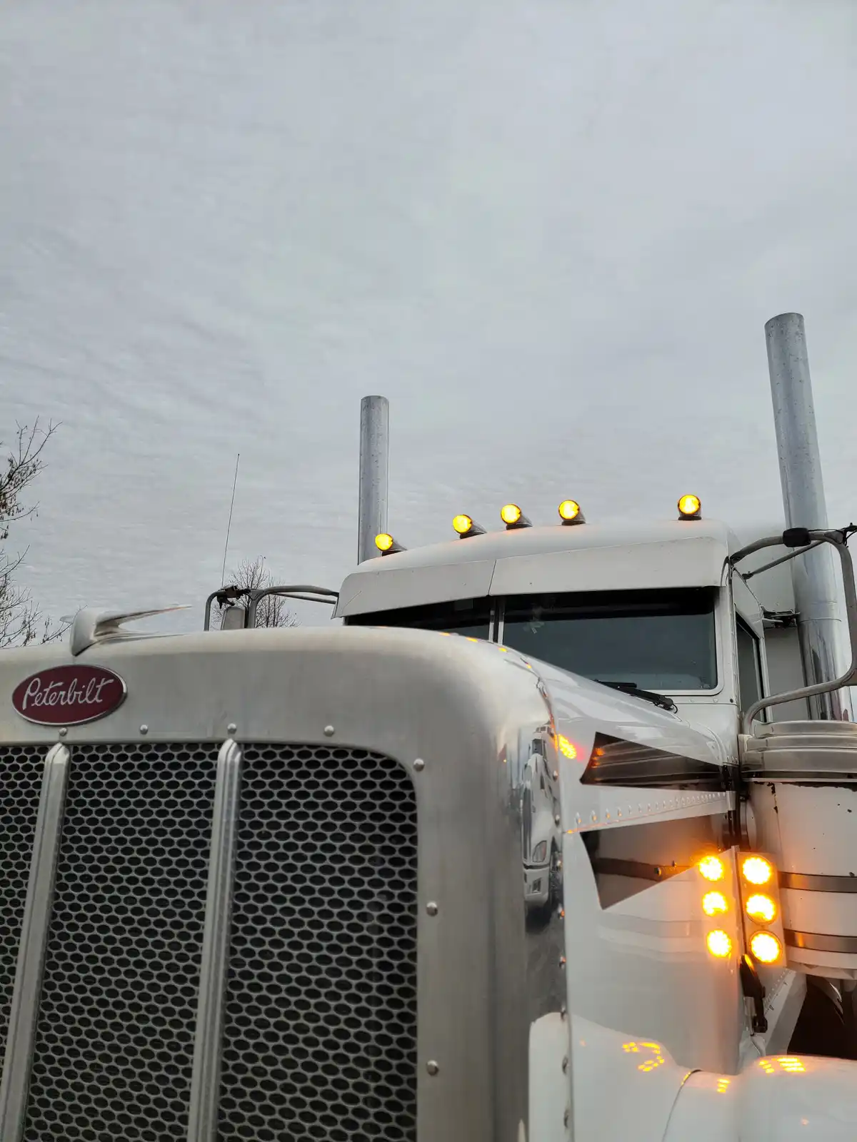 A low-angle shot of a white Peterbilt semi-truck with its amber lights on, set against an overcast sky. The truck's grille…