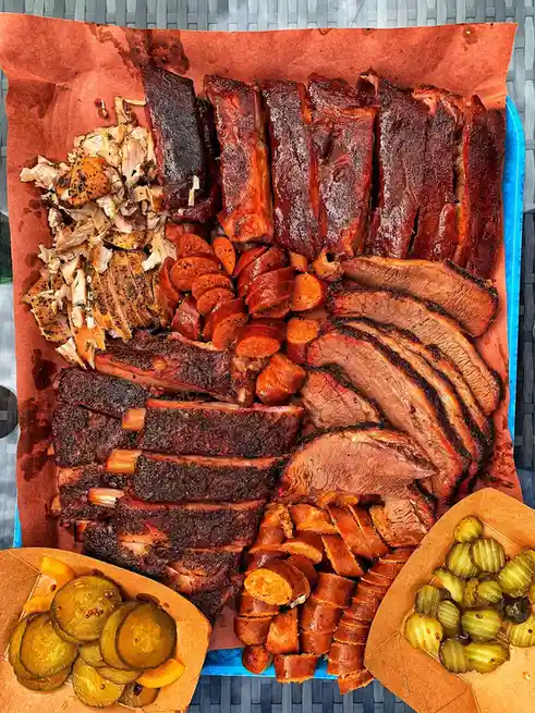 An overhead shot of a large blue tray lined with brown paper, overflowing with various smoked meats including ribs, brisket…