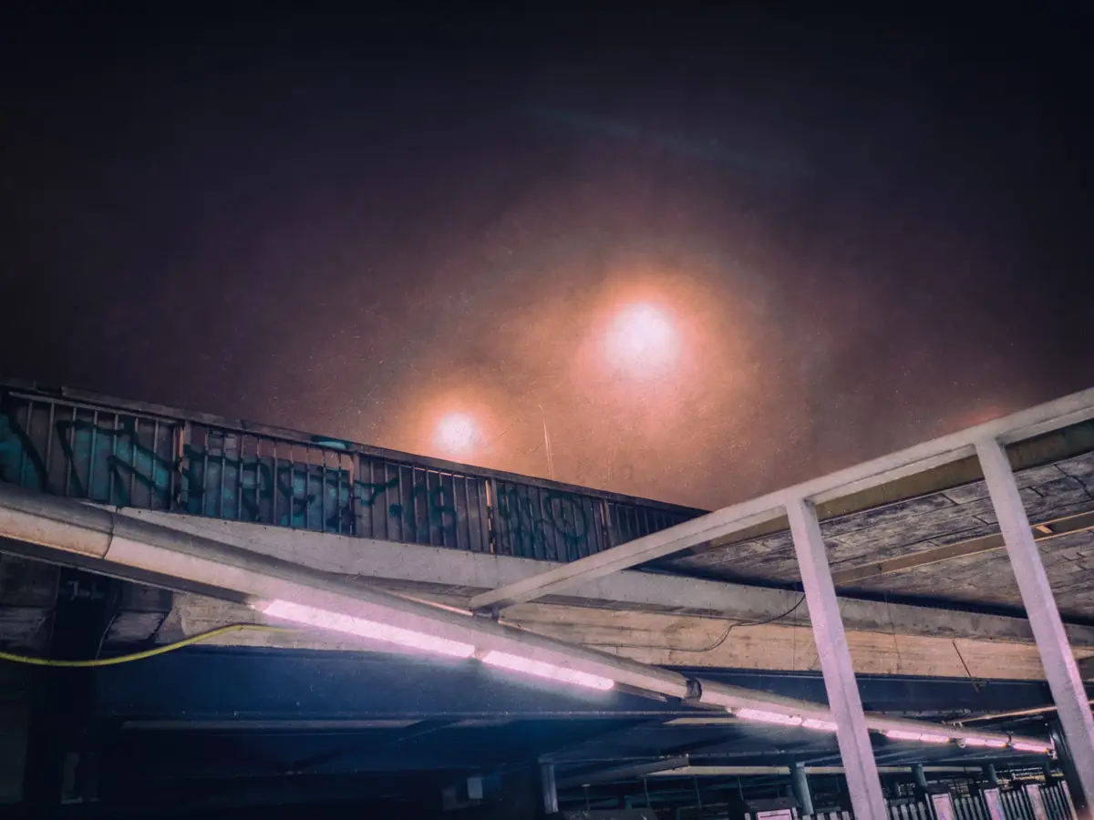 A low-angle nighttime photo of an urban overpass or parking structure, showing concrete beams, metal railings with graffiti…