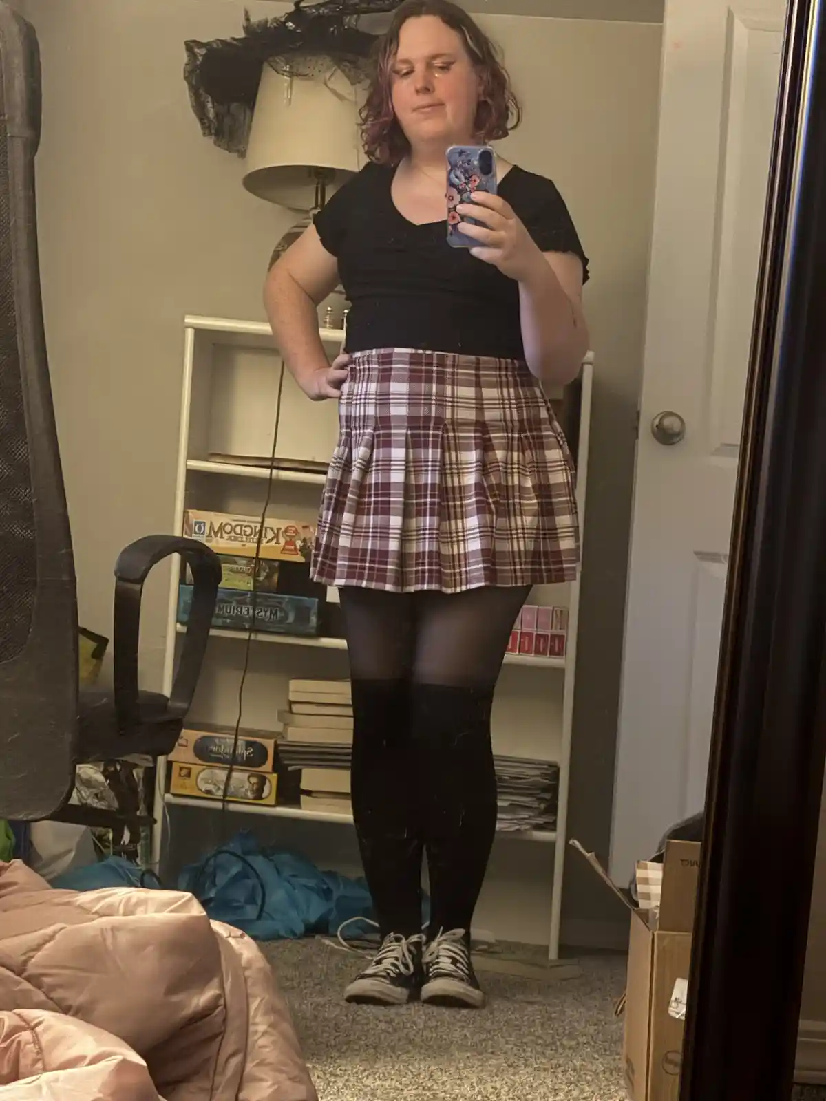 A person stands in front of a mirror in a bedroom, taking a selfie with a phone. They are wearing a black top, a plaid…