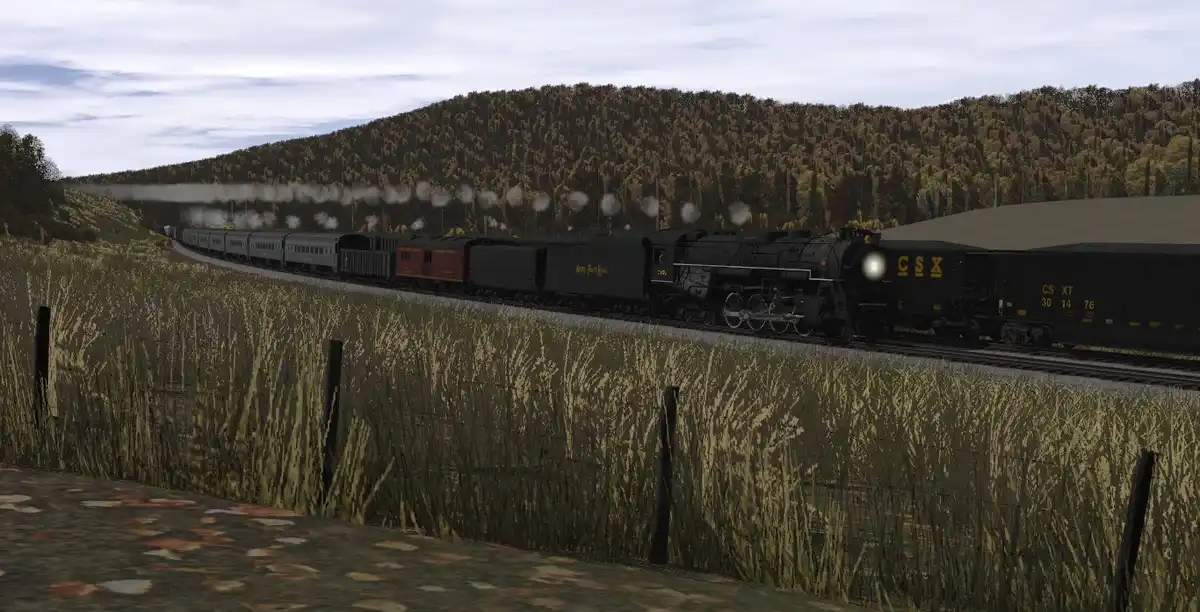 A steam locomotive pulling a long freight train travels along a track through a grassy field with a forested hill in the…