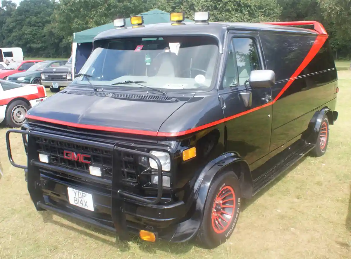 a black gmc van with bold red stripes running along its sides and roof rack, parked on grass at what appears to be a car…
