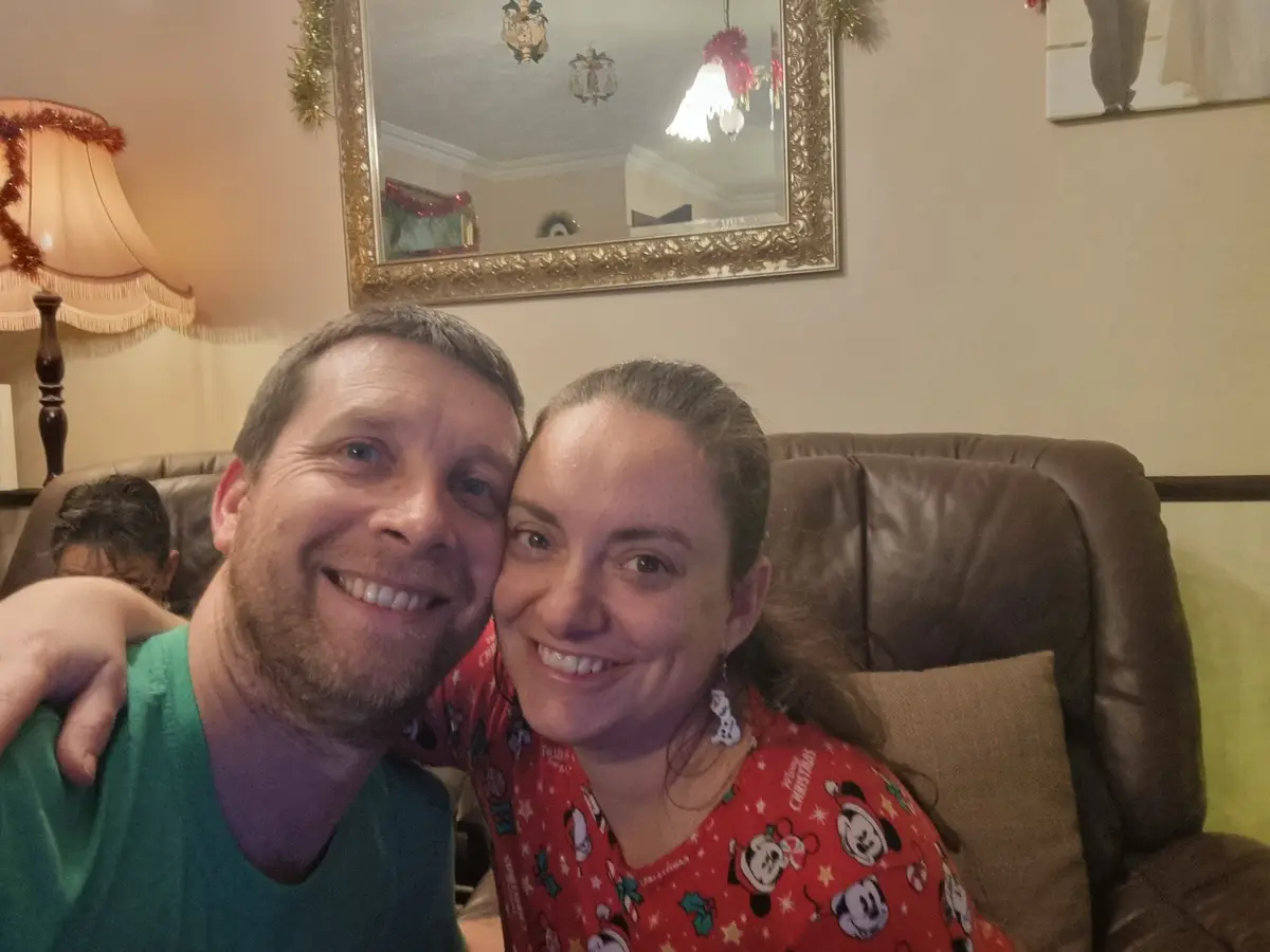 A man and woman are smiling closely together on a brown leather couch in a warmly lit living room decorated for Christmas…
