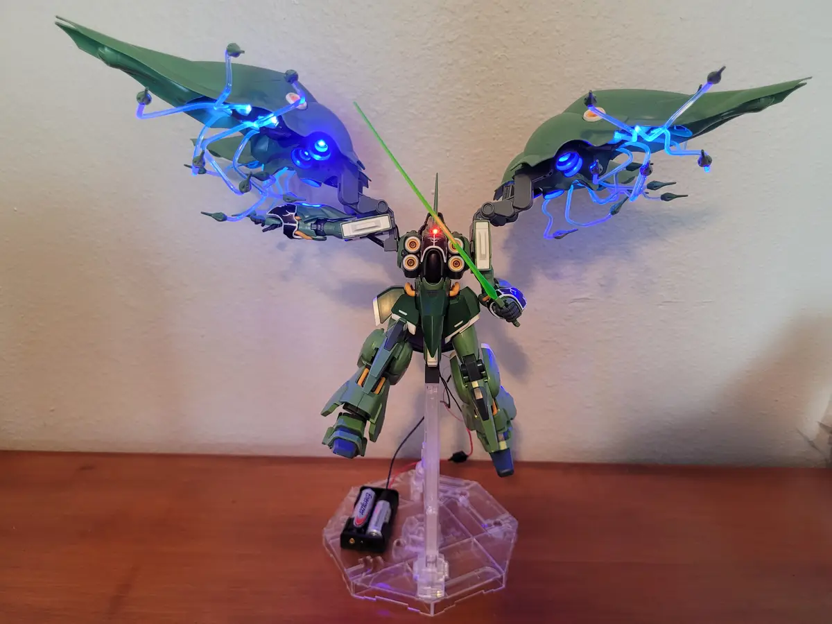 A green mecha model with glowing blue wings and a green beam saber stands on a clear plastic stand with batteries…