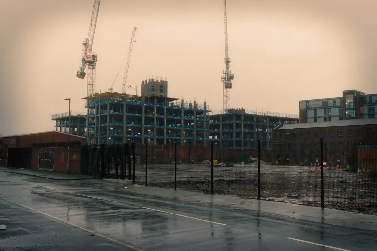 A photo of a large, multi-story building under construction with cranes and scaffolding, viewed from across a wet street…