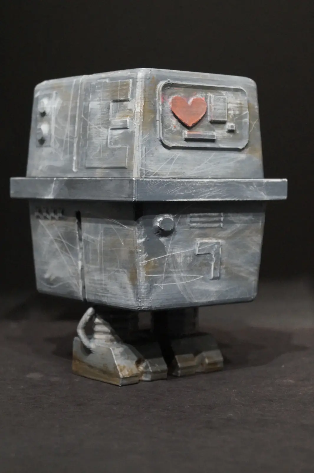 A small, boxy, metallic robot figurine with a worn, scratched surface stands against a dark background. Its faceplate…