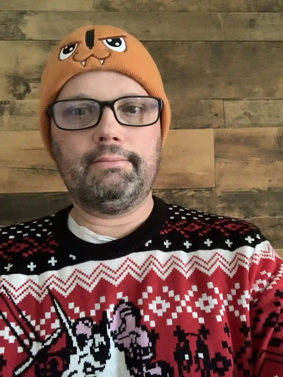 A man with glasses and a graying beard wears an orange beanie with a cartoon monster face and a red, black, and white…