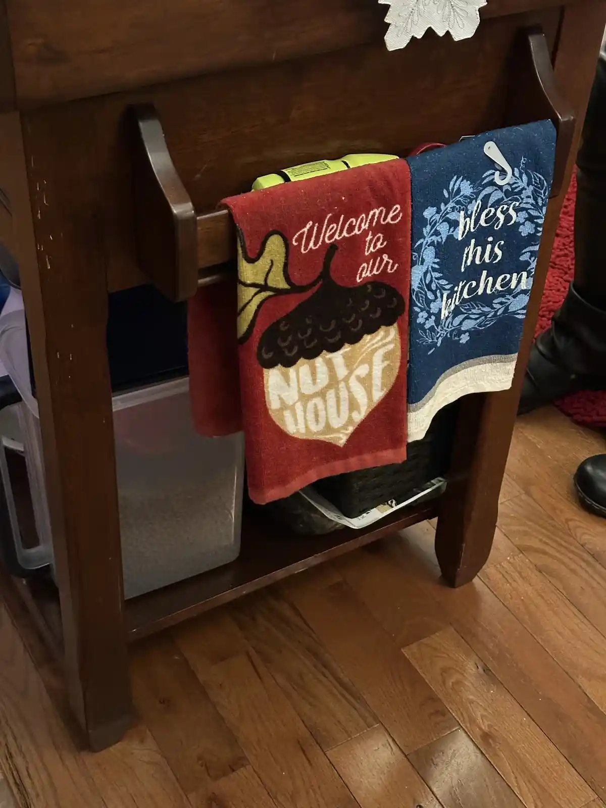 Two kitchen towels hang on a wooden table rack; one red towel reads 'Welcome to our nut house' with an acorn graphic, and a…