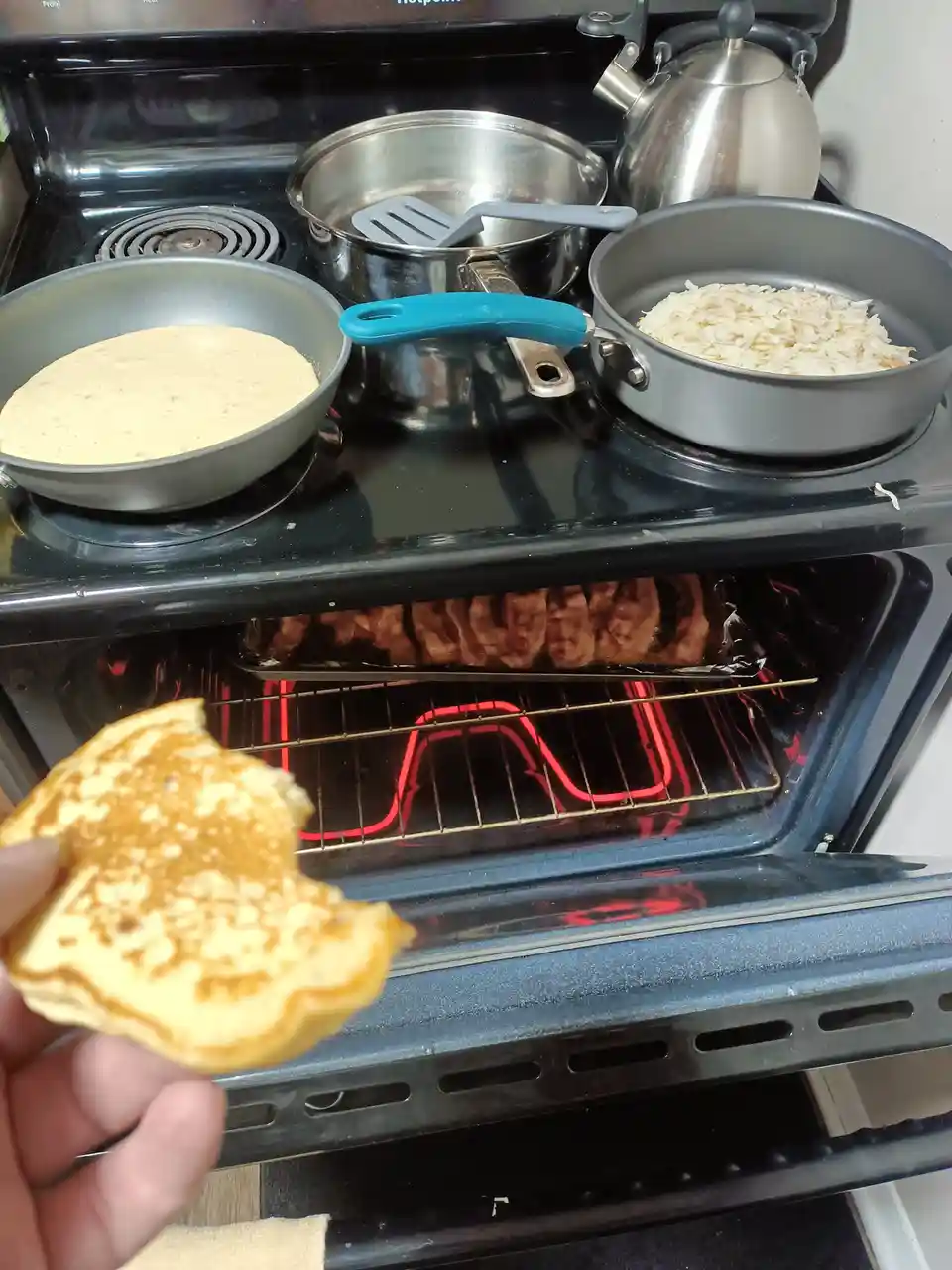 A hand holds a partially eaten pancake in front of an open oven with bacon cooking on the rack. The stovetop behind has a…