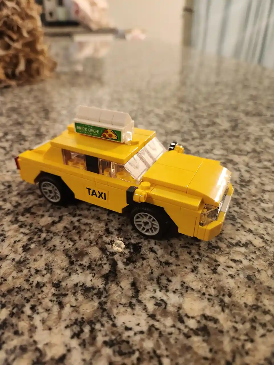 A yellow LEGO model of a taxi cab sits on a speckled countertop, featuring 'TAXI' printed on its side and a rooftop sign…