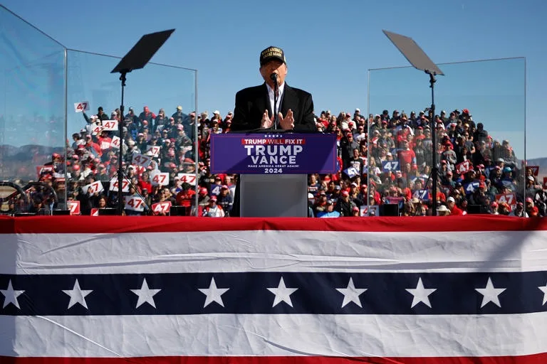 A man in a black suit and 'MAKE AMERICA GREAT AGAIN' cap stands at a podium with a blue banner reading 'TRUMP WILL FIX IT…