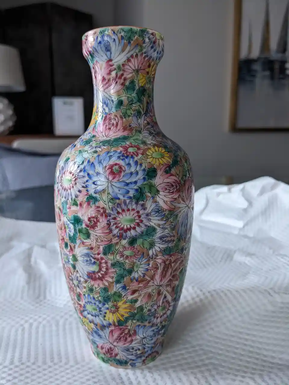 A colorful, hand-painted ceramic vase covered in dense floral patterns sits on a white textured surface. The vase features a…