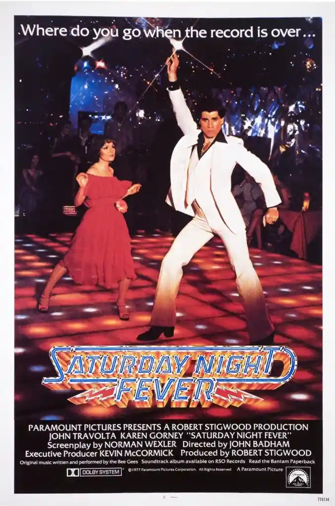 A vintage movie poster for 'Saturday Night Fever' shows John Travolta in a white suit dancing energetically on a disco floor…