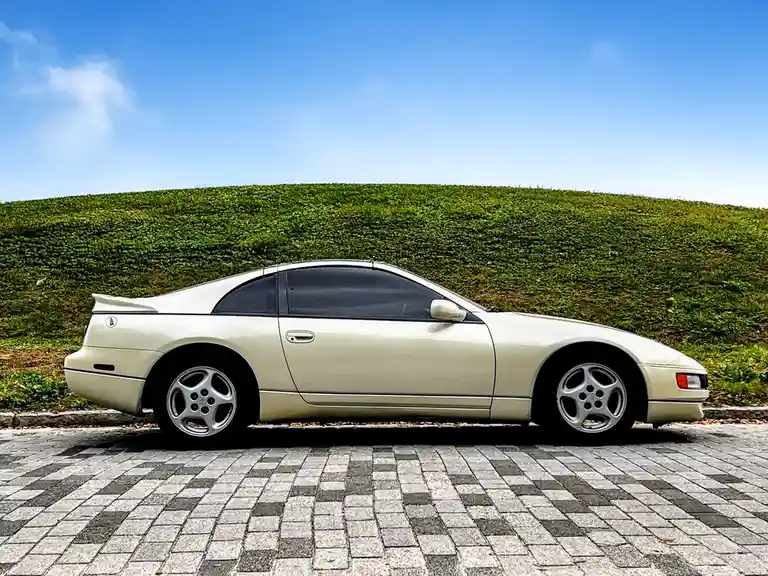 A cream-colored Nissan 300ZX sports car is parked on a patterned brick road in front of a green grassy hill under a blue…