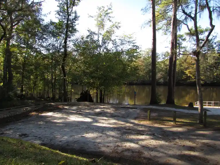 An empty, paved parking spot next to a calm body of water surrounded by trees, with two blue handicap parking signs visible…